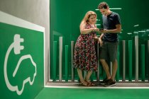 Sorrindo casal alegre à espera de carro elétrico para carregar e compartilhar telefone celular enquanto está em pé no salão de luz — Fotografia de Stock