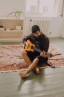 Уважний чоловічий музикант грає на гітарі, сидячи босоніж на підлозі сучасної квартири — стокове фото