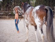 Side view of calm kid wearing feather Indian war bonnet and walking shirtless on sandy farm, leading horse behind — Stock Photo