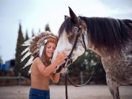 Enfant tranquille aux yeux fermés portant un bonnet de guerre indien traditionnel, collant avec un étalon de cheval sur un fond flou — Photo de stock