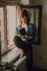 Vue de face de la femme détendue en tenue décontractée livre de lecture tout en étant assis pieds nus sur le rebord de la fenêtre dans l'appartement — Photo de stock