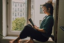 Side view of relaxed woman in casual outfit reading book while sitting barefoot on window sill in apartment — Stock Photo