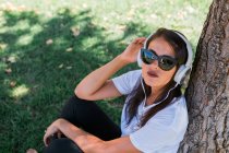 Peaceful woman in sunglasses and headphone listening to music while sitting on grass in park — Stock Photo