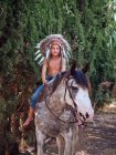Happy boy in authentic Indian feather hat riding horse in park — Stock Photo