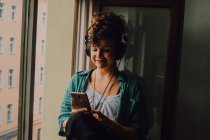 Mujer rizada sonriente en auriculares escuchando música mientras navega por el teléfono inteligente y se sienta en el alféizar de la ventana en el apartamento - foto de stock