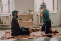 Side view of hipster woman taking picture of man sitting barefoot and posing on apartment floor — Stock Photo