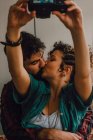 Cariñosa hipster pareja besándose y tomando selfie en cámara mientras sentado en piso en casa - foto de stock