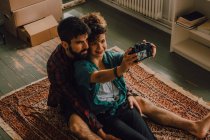 Сверху вид ласковой хипстерской пары, обнимающей и делающей селфи с фотокамерой, сидящей на полу дома — стоковое фото