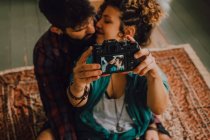Сверху вид ласковой хипстерской пары целующейся и делающей селфи с фотокамерой, сидящей на полу дома — стоковое фото