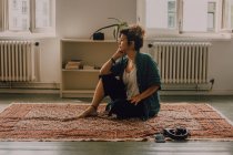 Расслабленная задумчивая женщина в повседневной одежде сидит на ковре в современной квартире — стоковое фото