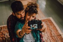 From above view of affectionate hipster couple hugging and taking selfie with photo camera while sitting on floor at home — Stock Photo