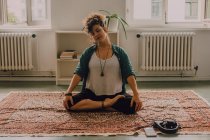Relaxed woman in casual clothes smiling and sitting in lotus pose on carpet in modern apartment — Stock Photo
