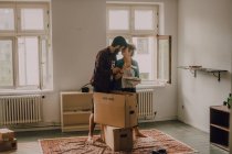 Пара хипстеров распаковывает вместе коробки, стоя босиком в светлой комнате и обнимаясь — стоковое фото