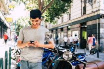 Positivo casual asiatico uomo utilizzando smartphone mentre in piedi in moto sulla strada soleggiata — Foto stock