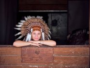 Delighted woman in traditional Indian feather war bonnet leaning on wooden fence on ranch — Stock Photo
