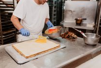 Булочник в латексных перчатках разбрасывает сладкое варенье на свежей булочке над кухонным прилавком — стоковое фото