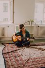 Розслаблена жінка в повсякденному одязі грає на акустичній гітарі, сидячи на підлозі в мінімалістичній кімнаті — стокове фото