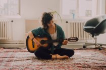 Donna rilassata in abiti casual suonare la chitarra acustica mentre seduto sul pavimento in camera minimalista — Foto stock