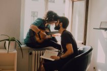 Счастливая женщина, играющая на гитаре, сидящая на подоконнике и целующийся мужчина с книгой — стоковое фото