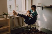 Relaxado mulher descalça em roupas casuais tocando guitarra acústica enquanto sentado na cadeira ao lado da janela em casa — Fotografia de Stock