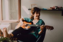 Donna rilassata a piedi nudi in abiti casual suonare la chitarra acustica mentre seduto sulla sedia accanto alla finestra a casa — Foto stock