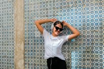 Hermosa mujer en traje casual y auriculares con las manos arriba junto a la pared de mosaico azul del edificio en la calle de la ciudad - foto de stock