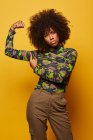 Mujer afroamericana fuerte y seria con camisa de camuflaje que muestra los biceps en el fondo amarillo mirando la cámara. - foto de stock