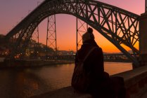 Vue arrière de touriste femelle regardant loin tout en étant assis sur la frontière de remblai de la ville près du pont pendant le coucher du soleil à Porto, Portugal — Photo de stock