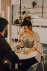 Радостная случайная женщина ест торт с вилкой, сидя за столом в кафетерии и глядя на своего парня — стоковое фото