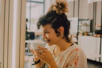 Vista lateral da mulher sorridente feliz sorvendo da caneca enquanto sentado na cafeteria moderna — Fotografia de Stock