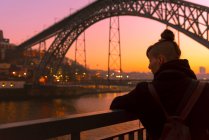 Вид сзади женщины-туристки, стоящей рядом с городской набережной возле моста, смотрящей на закат в Порту, Португалия — стоковое фото