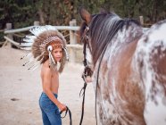 Ruhiges Kind mit Federhaube und ohne Hemd auf sandigem Bauernhof, führendes Pferd hinter sich — Stockfoto