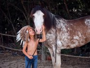Delighted kid in Indian feather war bonnet caressing horse on ranch and looking away — Stock Photo