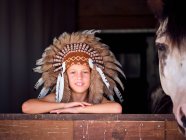 Enfant enchanté dans un authentique chapeau de plume indien appuyé sur une clôture en bois dans une écurie avec cheval sur un ranch et regardant la caméra — Photo de stock