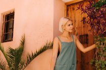 Belle jeune femme aux cheveux blonds courts regardant loin et s'appuyant sur le mur tout en se tenant debout sur une vieille porte en bois — Photo de stock