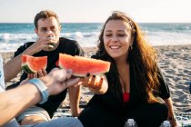 Улыбающаяся женщина дает кусочек арбуза своей подруге, которая пьет апельсиновый сок на пляже — стоковое фото