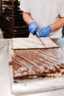 Обрізати людину в уніформі і рукавичках за допомогою ножа, щоб нарізати солодкий свіжий торт на столі в пекарні — стокове фото