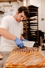 Кондитер в перчатках украшает вкусный свежий торт шоколадными брызгами во время работы в пекарне — стоковое фото