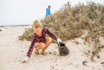 Positive Hipster-Frau in lässiger Kleidung und Handschuhen sammelt Müll in Tüten, während sie am menschenleeren Strand hockt — Stockfoto