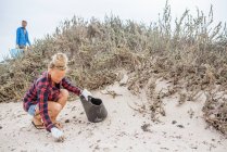 Positive hipster woman in casual clothes and gloves collecting trash into bag while squatting on deserted beach — Stock Photo