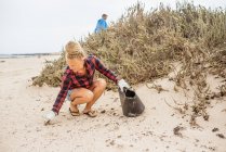 Positivo hipster donna in abiti casual e guanti raccolta spazzatura in borsa mentre accovacciato sulla spiaggia deserta — Foto stock