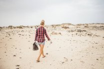 Positive Hipster-Frau in lässiger Kleidung und Handschuhen sammelt Müll in Tüten, während sie am menschenleeren Strand hockt — Stockfoto