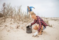 Positive hipster woman and man in casual clothes and gloves collecting trash into bag while squatting on deserted beach — Stock Photo