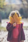 Little girl in casual outfit hiding face behind yellow maple leaf while standing on sunny autumn daytime in park — Stock Photo