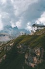 Edifício branco e cinza na colina rochosa com grama grossa verde contra belas montanhas de névoa em Dolomitas durante o tempo nublado — Fotografia de Stock