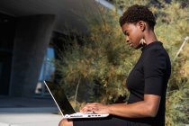 Side view of concentrated African American woman in elegant black dress using laptop on street — Stock Photo
