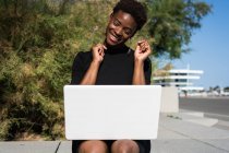 Cheerful excited woman in elegant black dress using laptop on pavement on street — Stock Photo