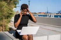 Разочарованная афроамериканка в черном платье с ноутбуком и разговаривающая по смартфону на улице — стоковое фото