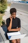 Mulher afro-americana decepcionada em vestido preto usando laptop e falando no smartphone na rua — Fotografia de Stock