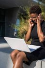 Афроамериканець жінка в елегантній чорній сукні з використанням ноутбука і виступаючи на мобільному телефоні на вулиці — стокове фото